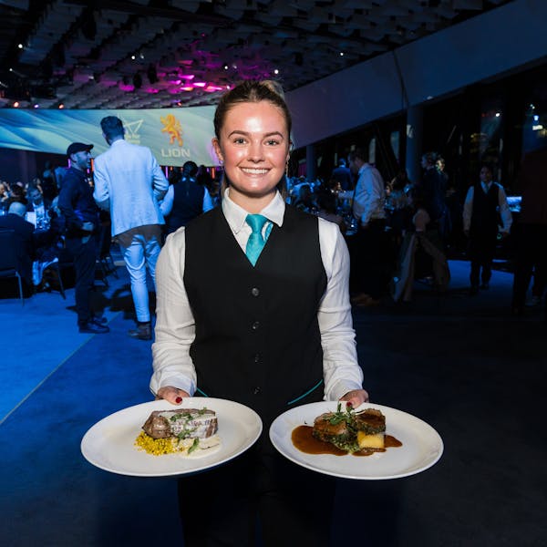 A server in formal attire presents two plates of food—one with a meat dish and the other decorated with vegetables—during a vibrant Te Pae event bustling with attendees.